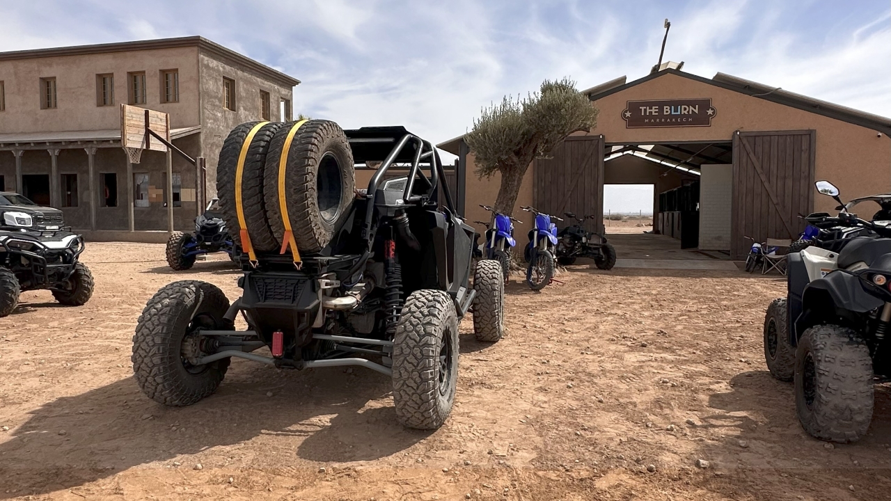 Off-road buggy with spare tyres parked on sandy ground in front of an adventure base